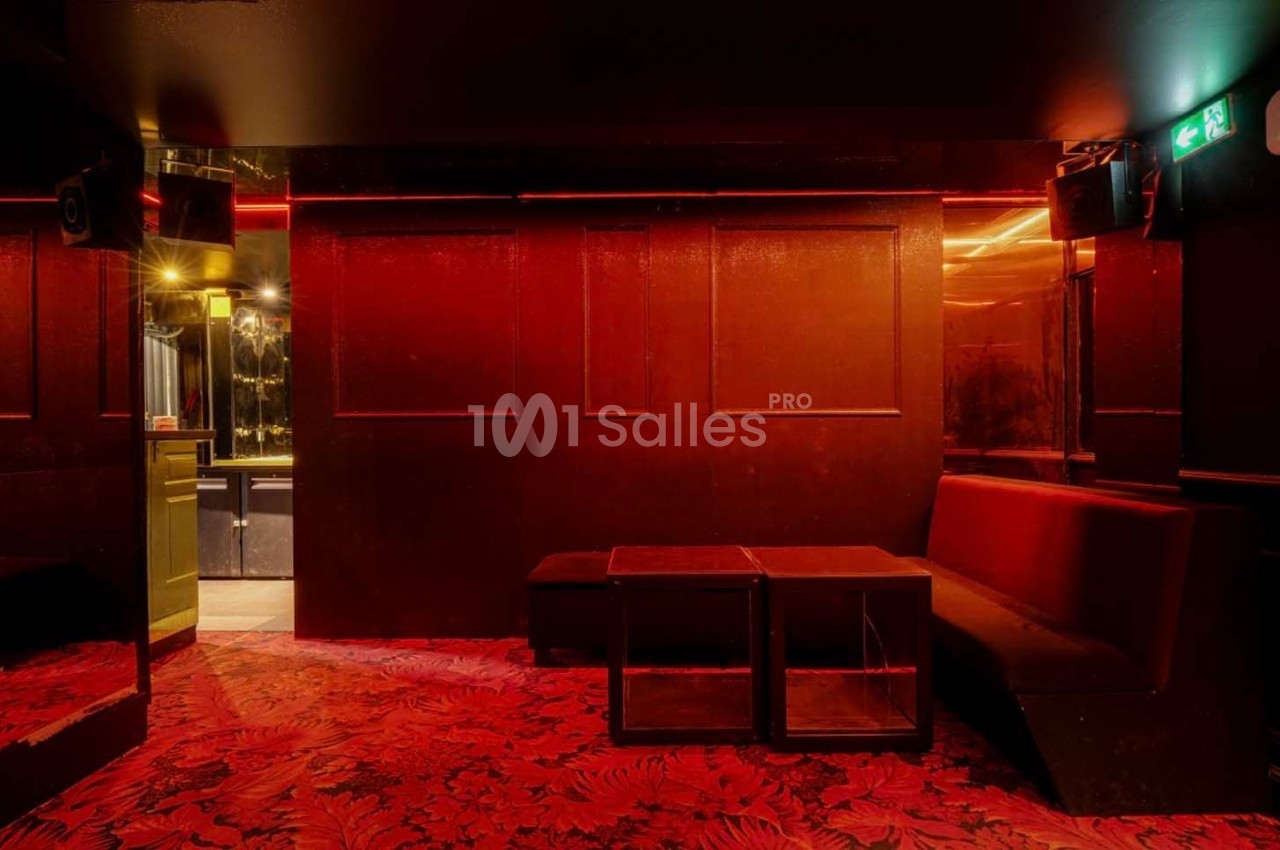 Salle sombre avec éclairage rouge, banquettes, tables basses et sol à motif végétal, entrée visible au fond.