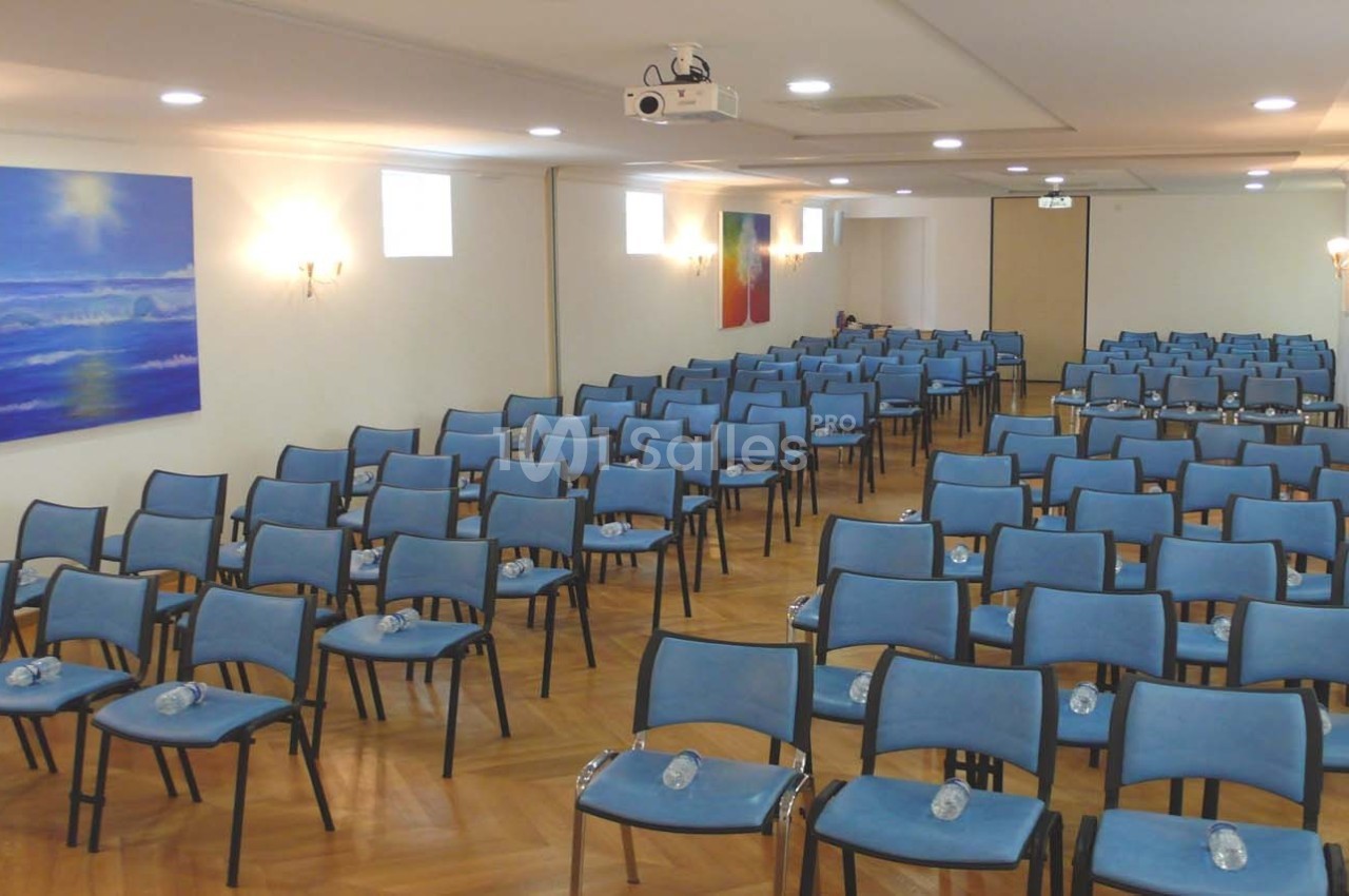 Salle de conférence lumineuse avec des rangées de chaises bleues alignées et des bouteilles d'eau posées sur chaque siège.