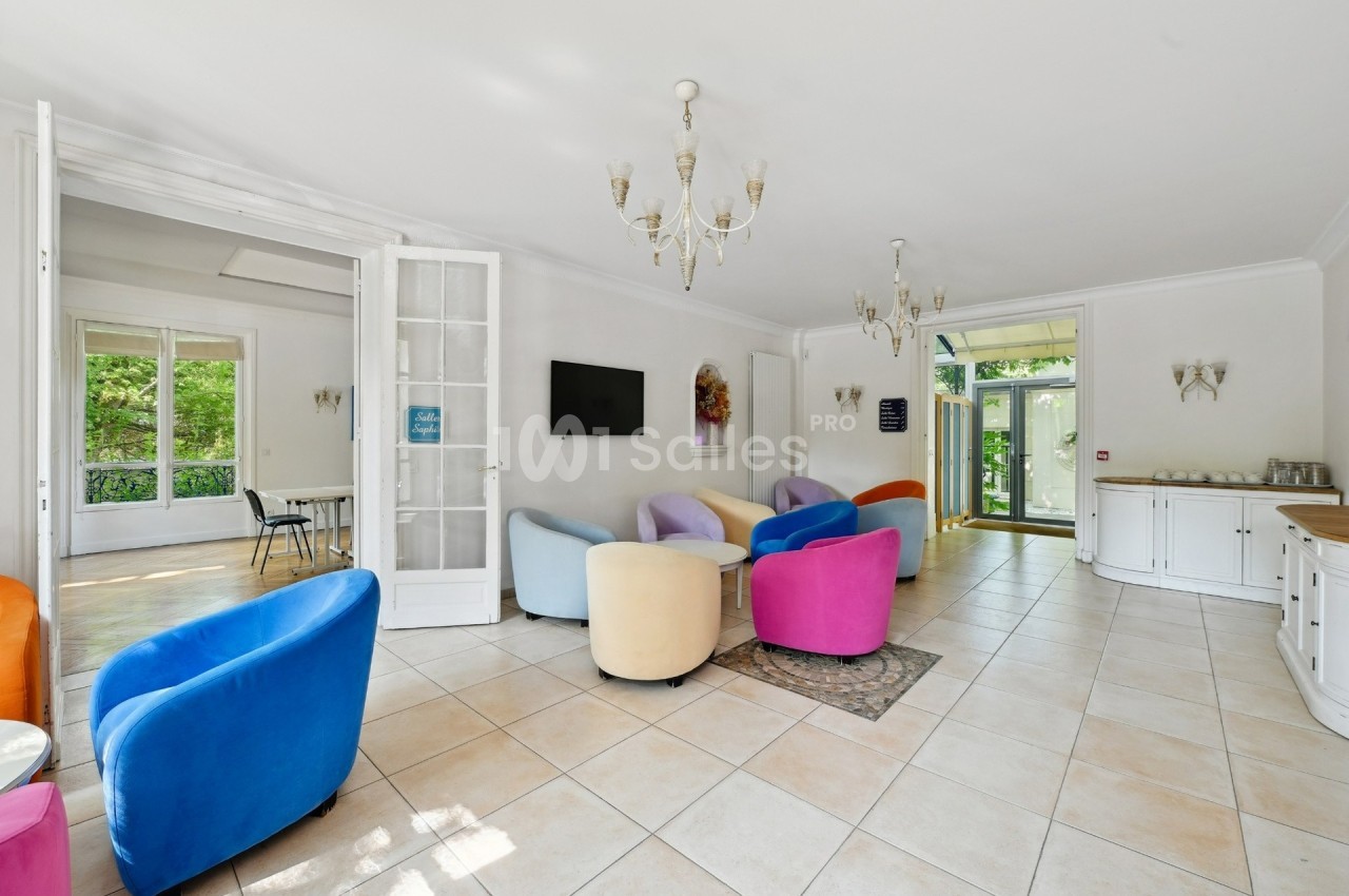 Salon lumineux avec fauteuils colorés, carrelage clair, lustres et vue sur un espace extérieur verdoyant.