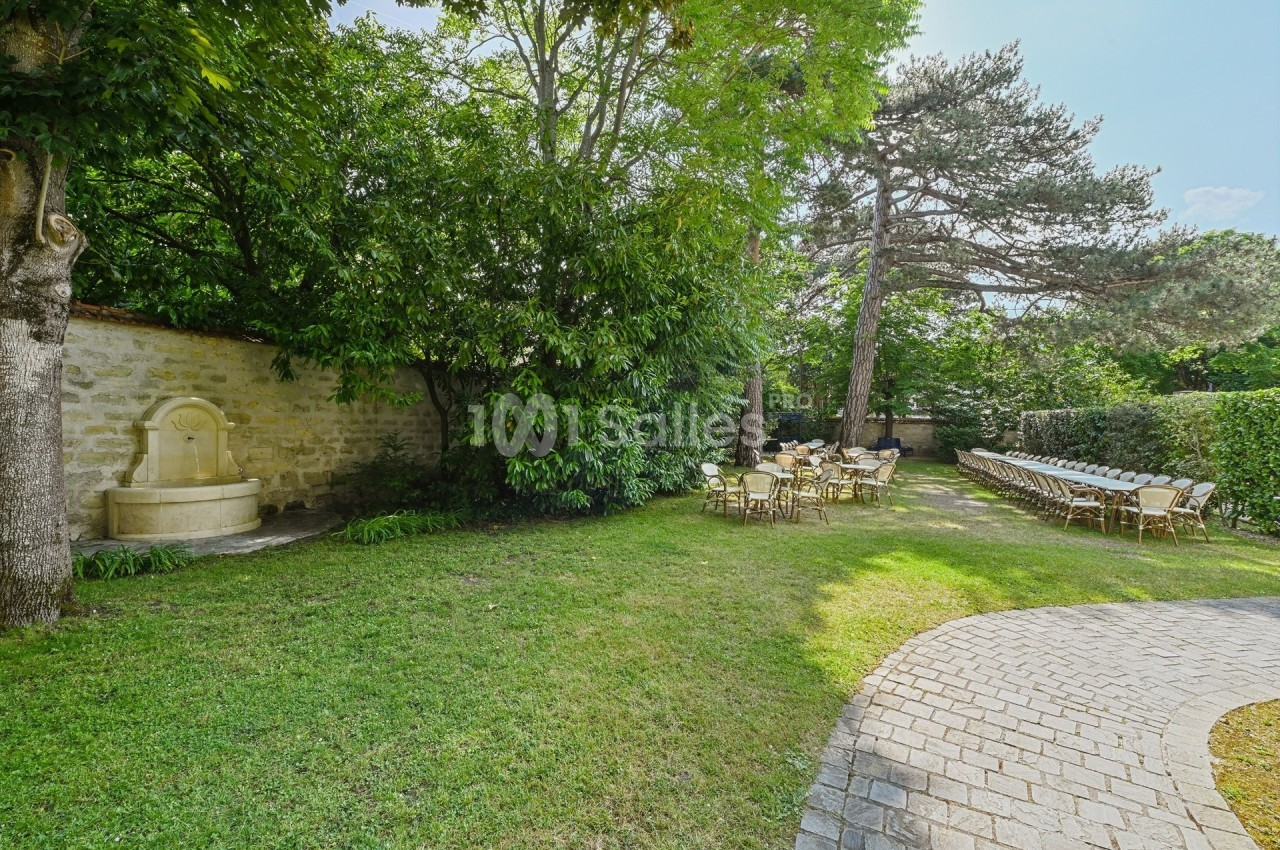 Jardin avec pelouse, fontaine en pierre, tables et chaises disposées sous des arbres près d'un chemin pavé.