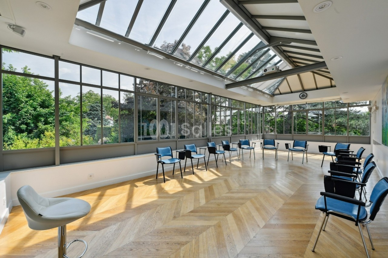 Salle lumineuse avec parquet en bois, grandes baies vitrées, chaises disposées en cercle et vue sur un jardin verdoyant.