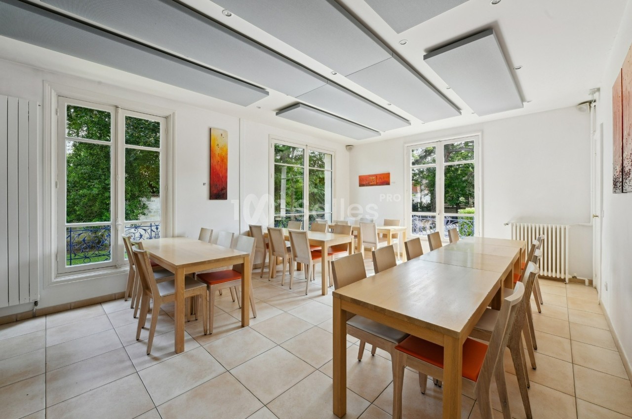 Salle lumineuse avec grandes fenêtres, tables en bois clair, chaises assorties et vue sur un jardin verdoyant.