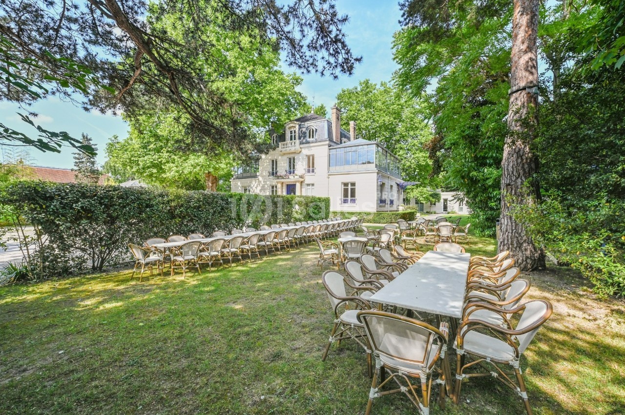 Tables et chaises disposées en extérieur dans un jardin verdoyant, avec une grande maison en arrière-plan.