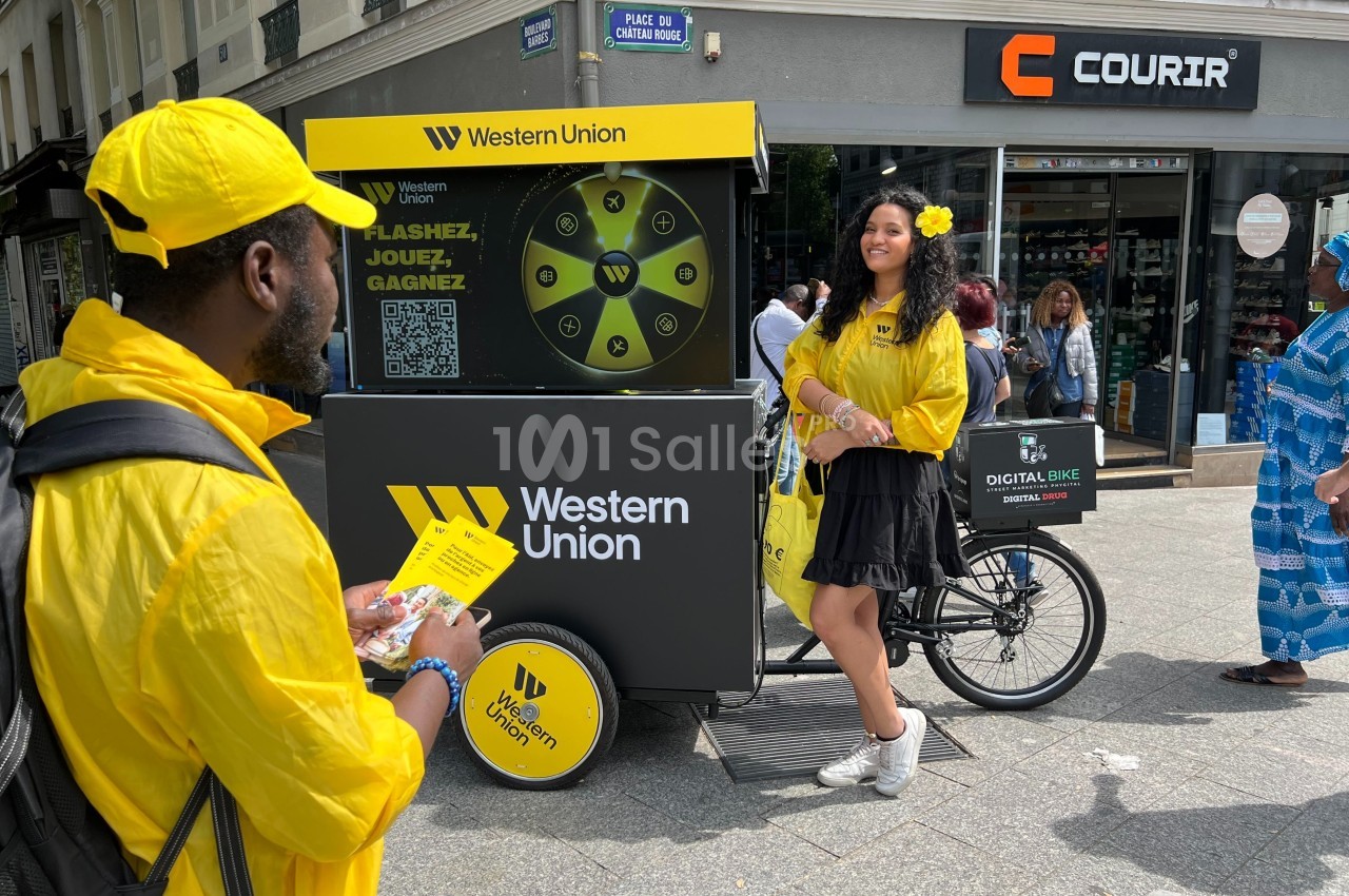 Deux personnes en tenue jaune près d'un vélo promotionnel Western Union dans une rue commerçante.