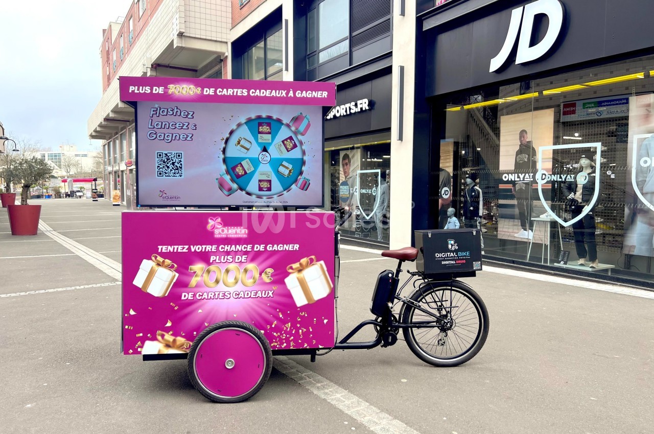 Triporteur promotionnel rose stationné devant des magasins, affichant une offre de cartes cadeaux à gagner.
