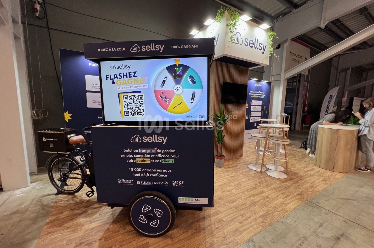 Stand d'exposition avec un écran interactif, une roue de loterie et un triporteur, dans un espace intérieur éclairé.