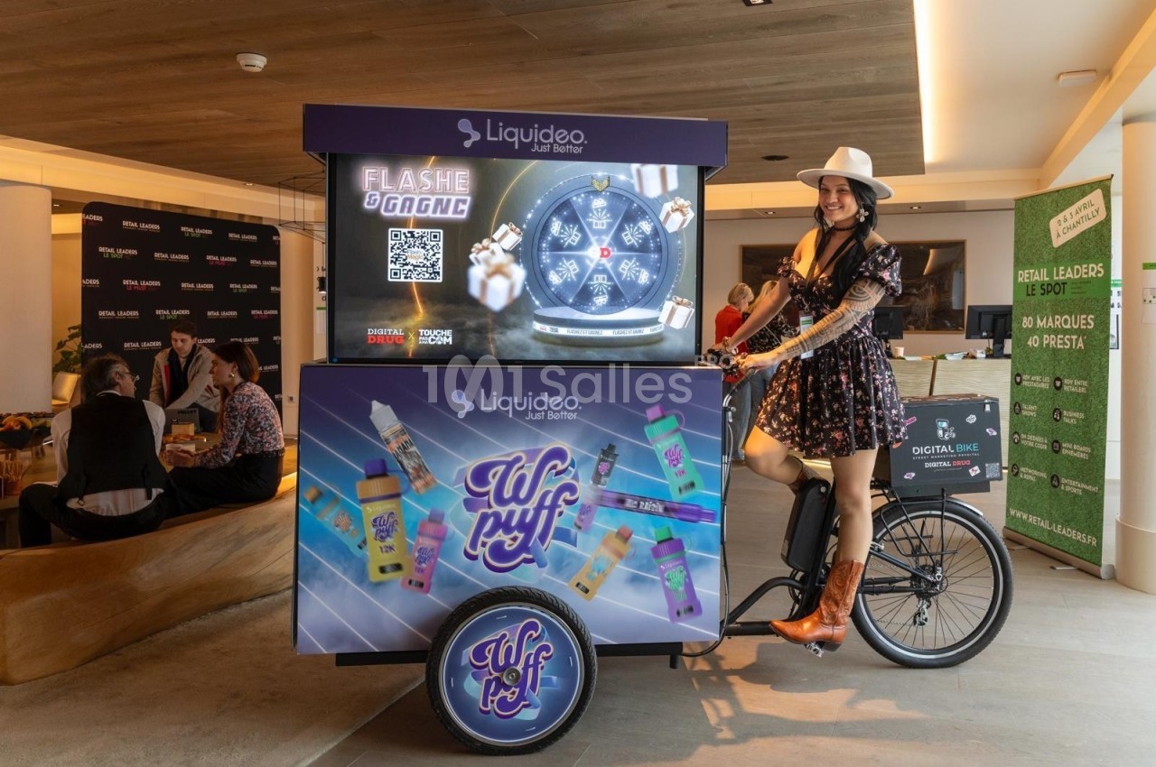 Une femme en tenue western pose près d'un triporteur promotionnel de produits de vape dans un espace intérieur moderne.