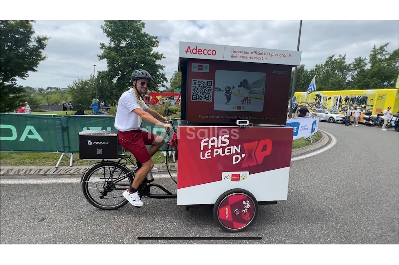 Un homme conduit un vélo cargo publicitaire Adecco sur une route bordée d'arbres et de spectateurs.