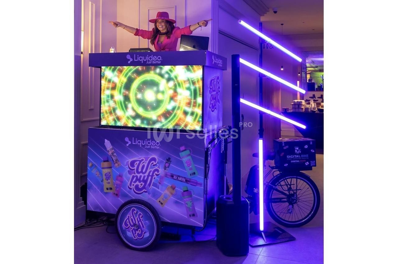 Femme souriante sur un stand lumineux décoré de motifs colorés, avec des tubes LED violets et un vélo intégré.