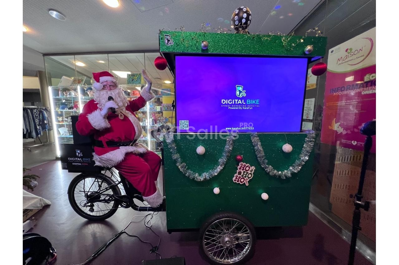 Un Père Noël assis sur un triporteur décoré, équipé d'un écran numérique affichant ’Digital Bike’.