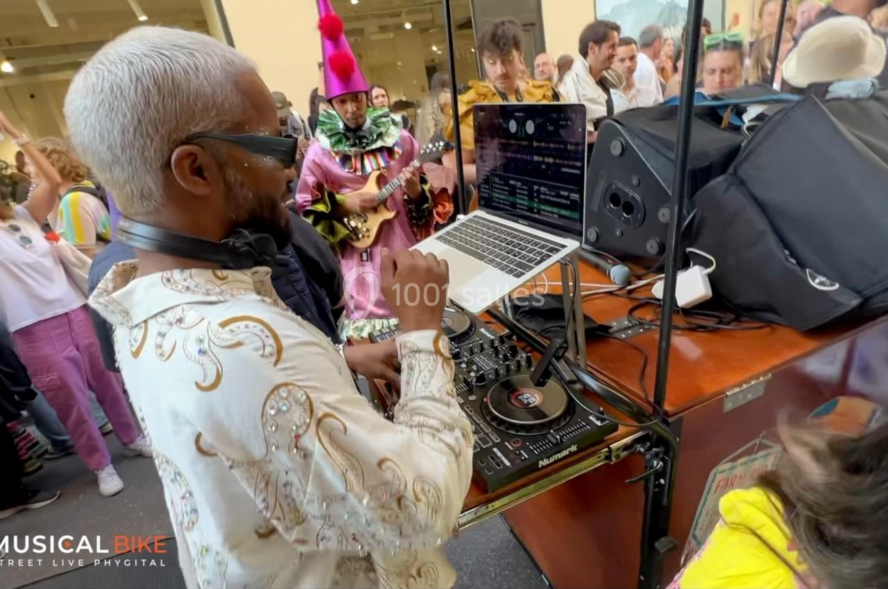 Un DJ mixe sur une table de mixage entouré de musiciens costumés et d'un public dans un espace animé.