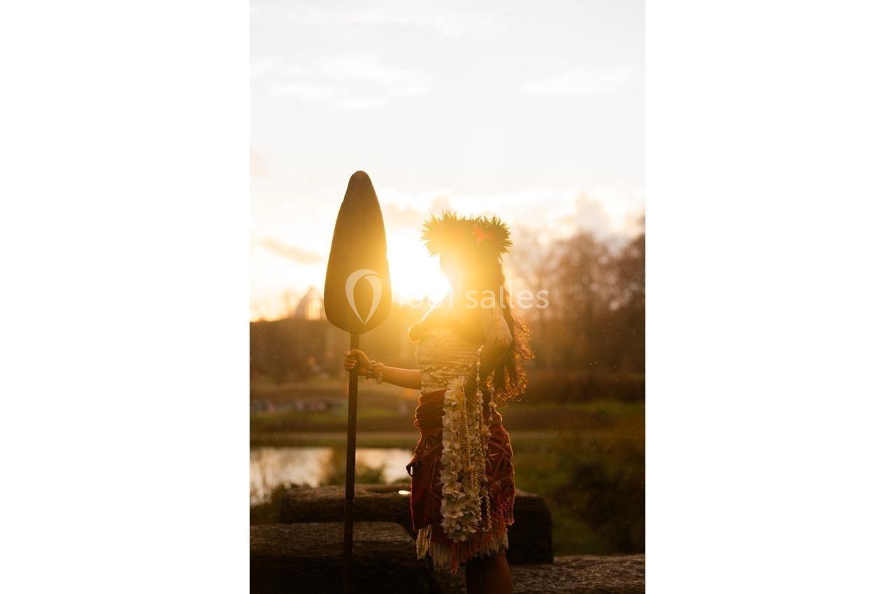 Personne en tenue traditionnelle polynésienne tenant une pagaie, éclairée par un coucher de soleil en arrière-plan.