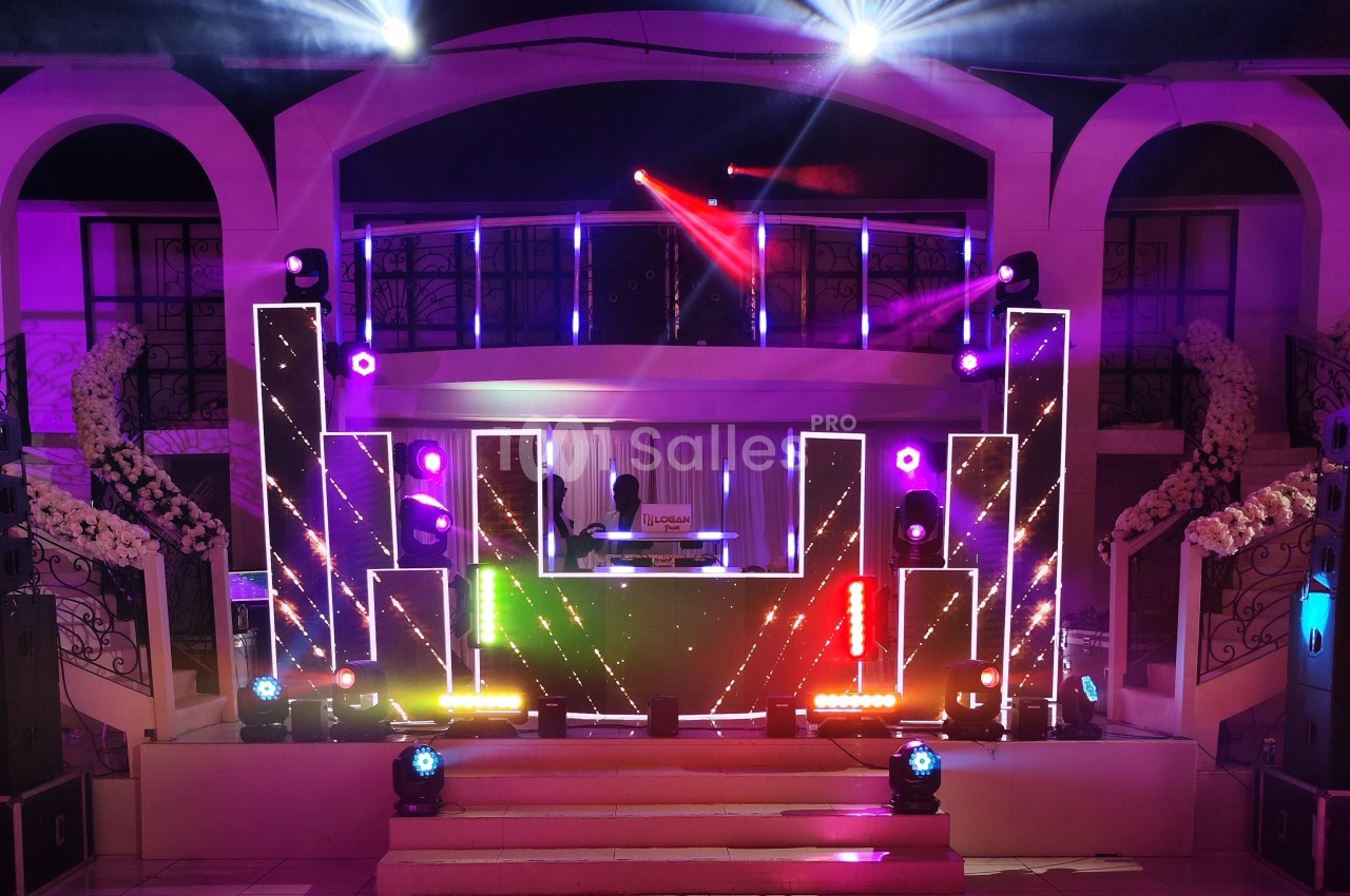Scène de concert éclairée avec un DJ aux platines, entourée de lumières colorées et d'une décoration élégante.