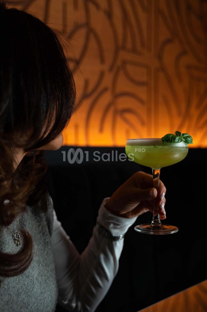 Une personne tenant un cocktail vert garni de feuilles de basilic dans un cadre tamisé.