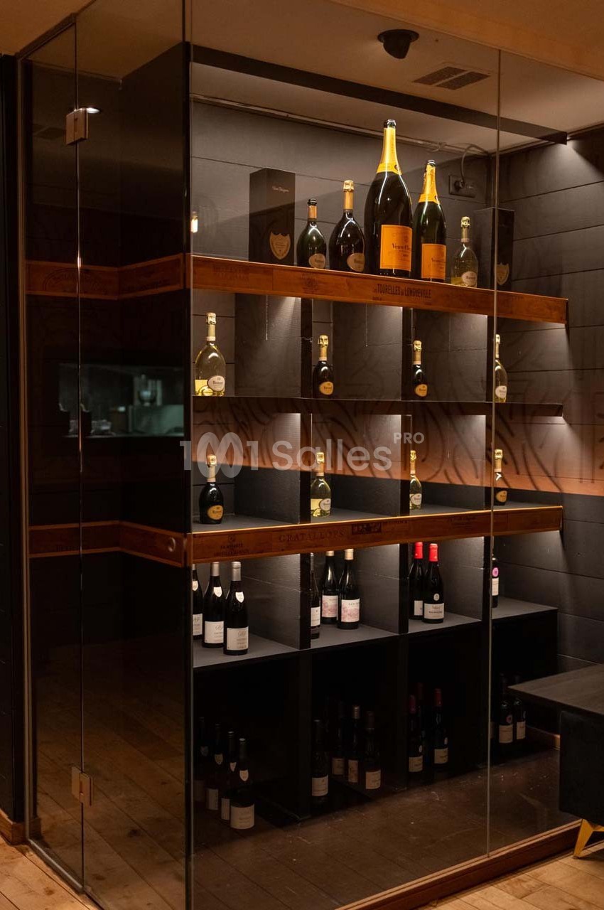 Cave à vin vitrée avec étagères en bois, contenant des bouteilles de vin et de champagne éclairées par des spots.