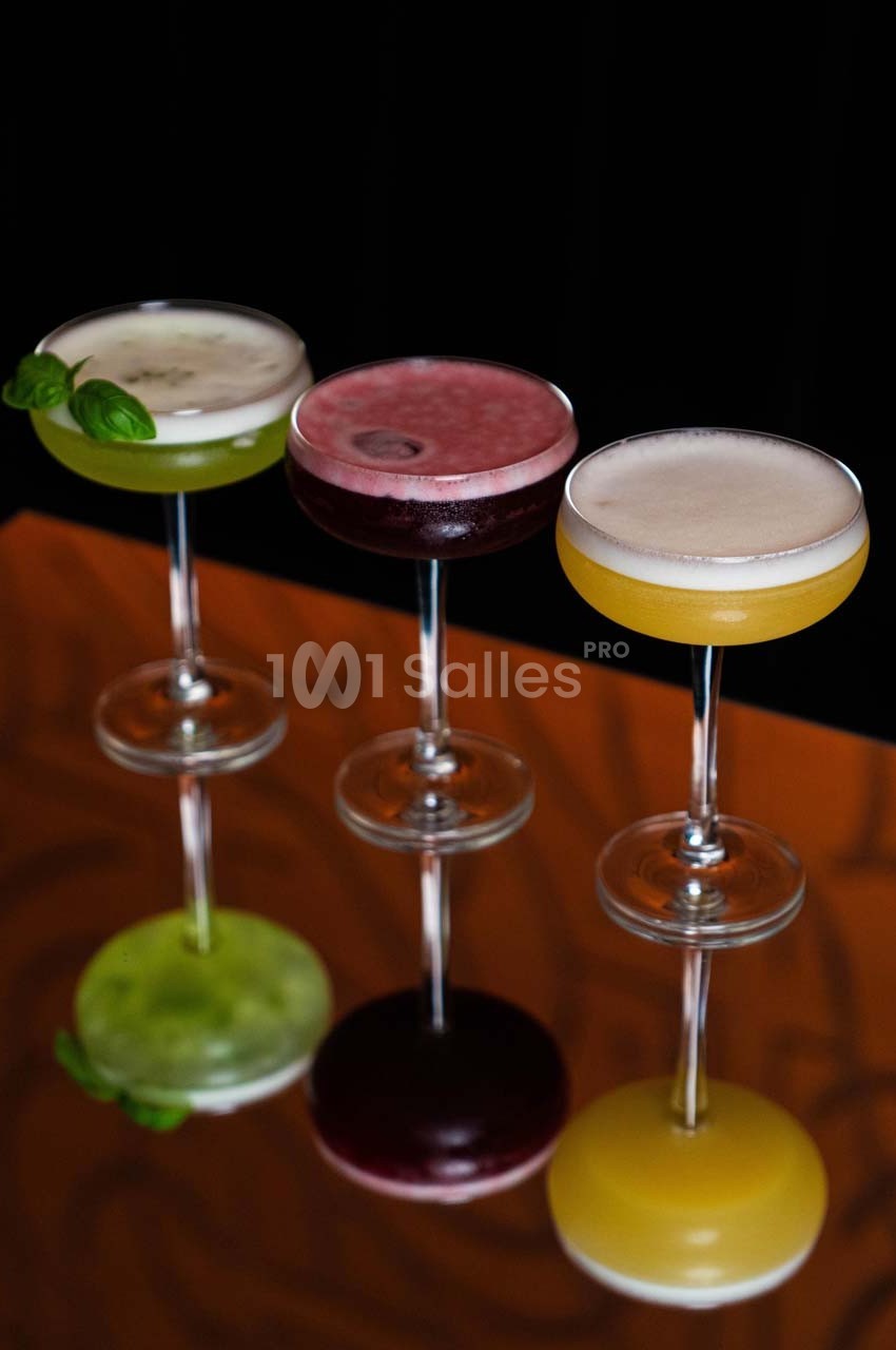 Trois cocktails colorés dans des verres à pied alignés sur une surface réfléchissante, fond sombre.