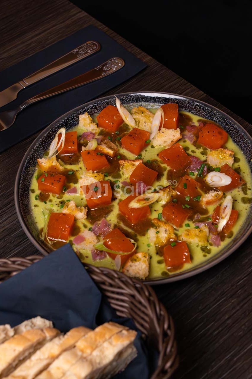 Assiette de cubes de potiron marinés avec sauce verte, oignons, croûtons et ciboulette, servie sur une table sombre.