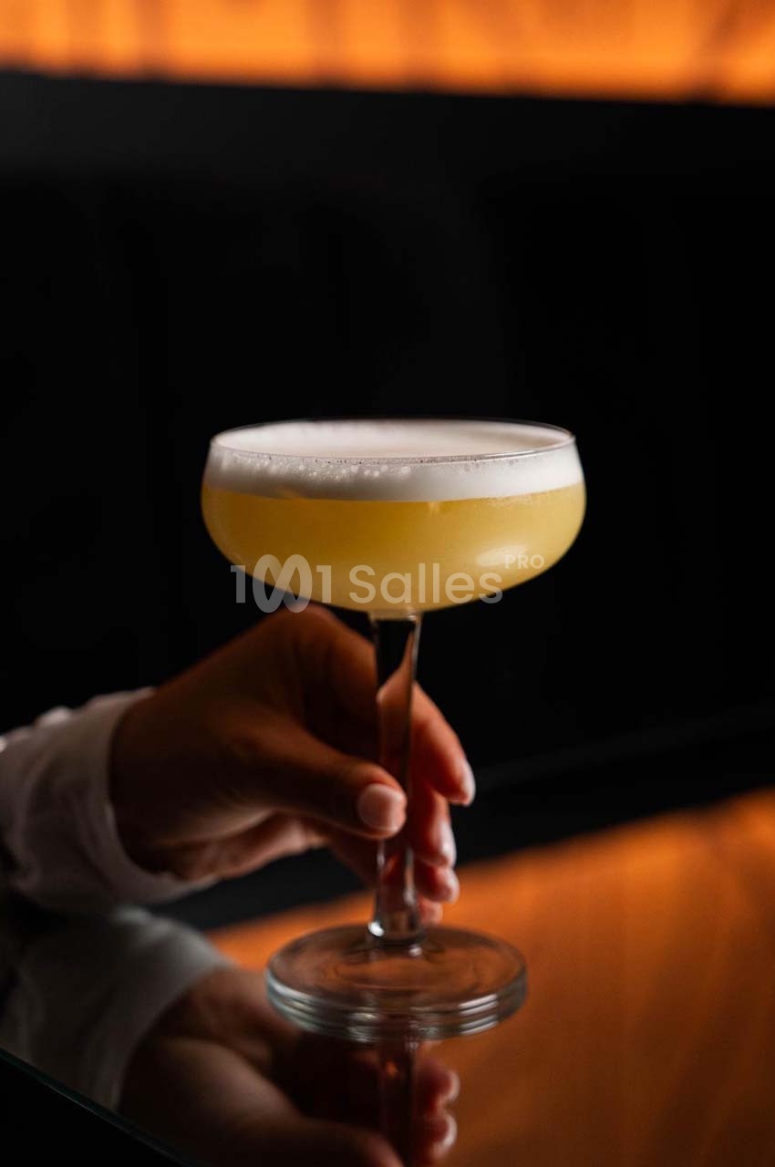Un verre à cocktail rempli d'une boisson mousseuse jaune pâle, tenu par une main sur une table sombre.