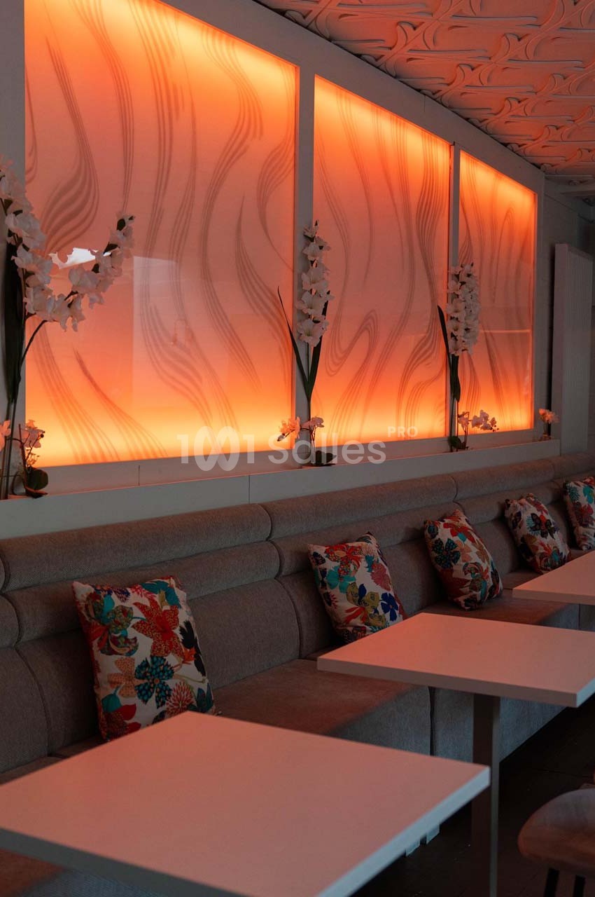 Banquettes grises avec coussins colorés alignées face à des tables blanches, éclairage mural orange en arrière-plan.