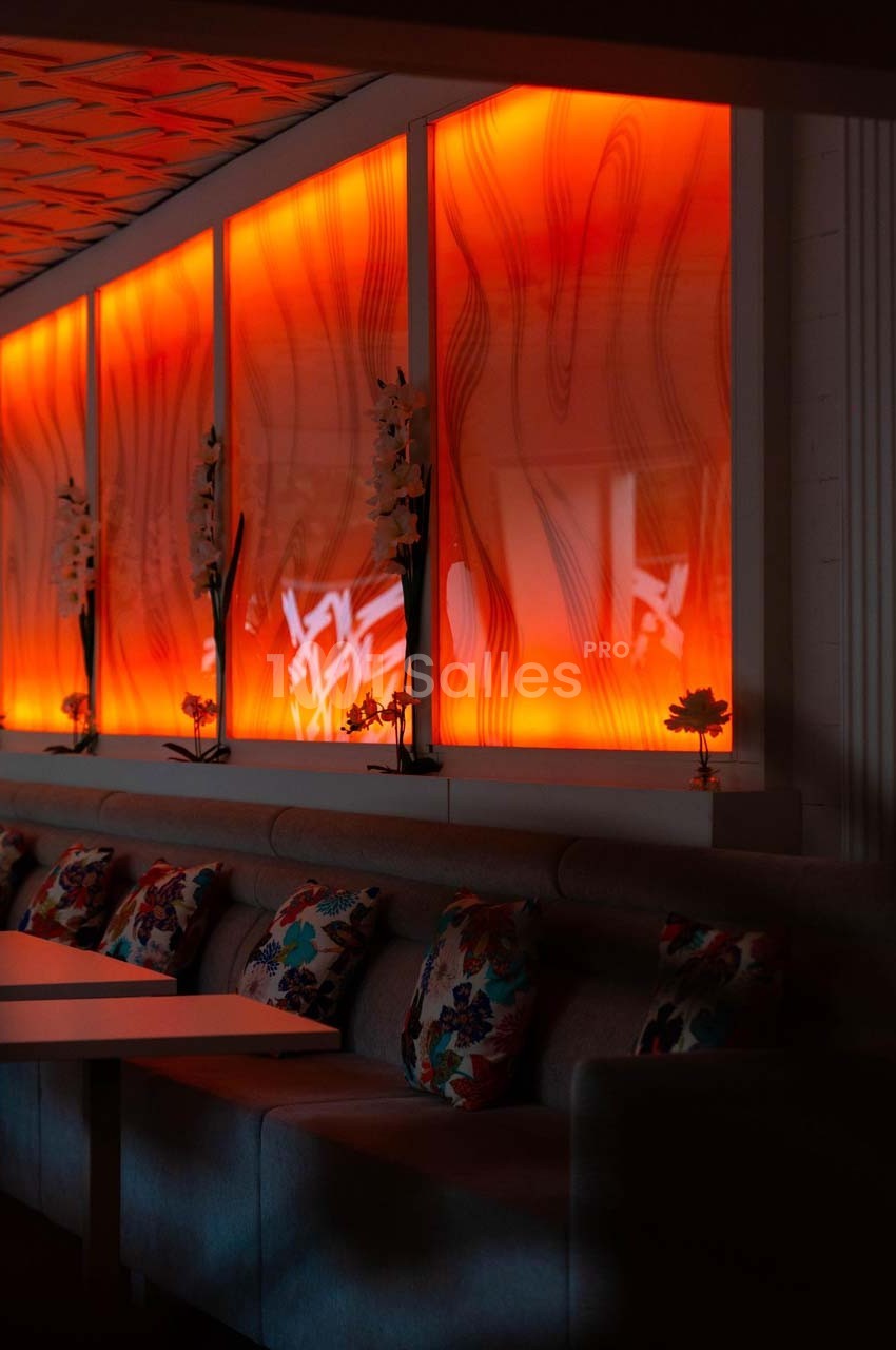 Mur rétroéclairé orange avec motifs ondulés, banquette grise ornée de coussins colorés et petites fleurs décoratives.