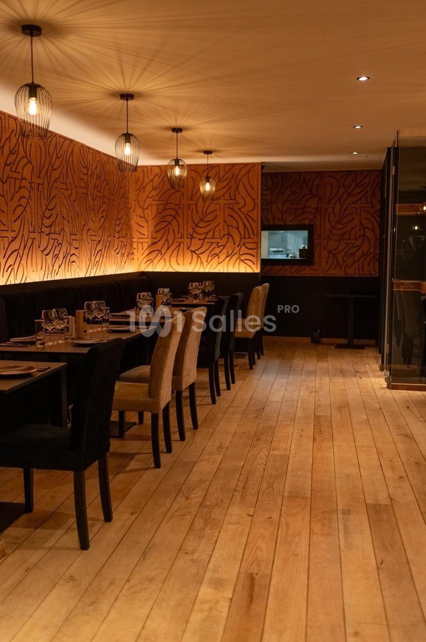 Salle de restaurant chaleureuse avec parquet en bois, murs ornés de motifs, et tables dressées sous des luminaires suspendus.