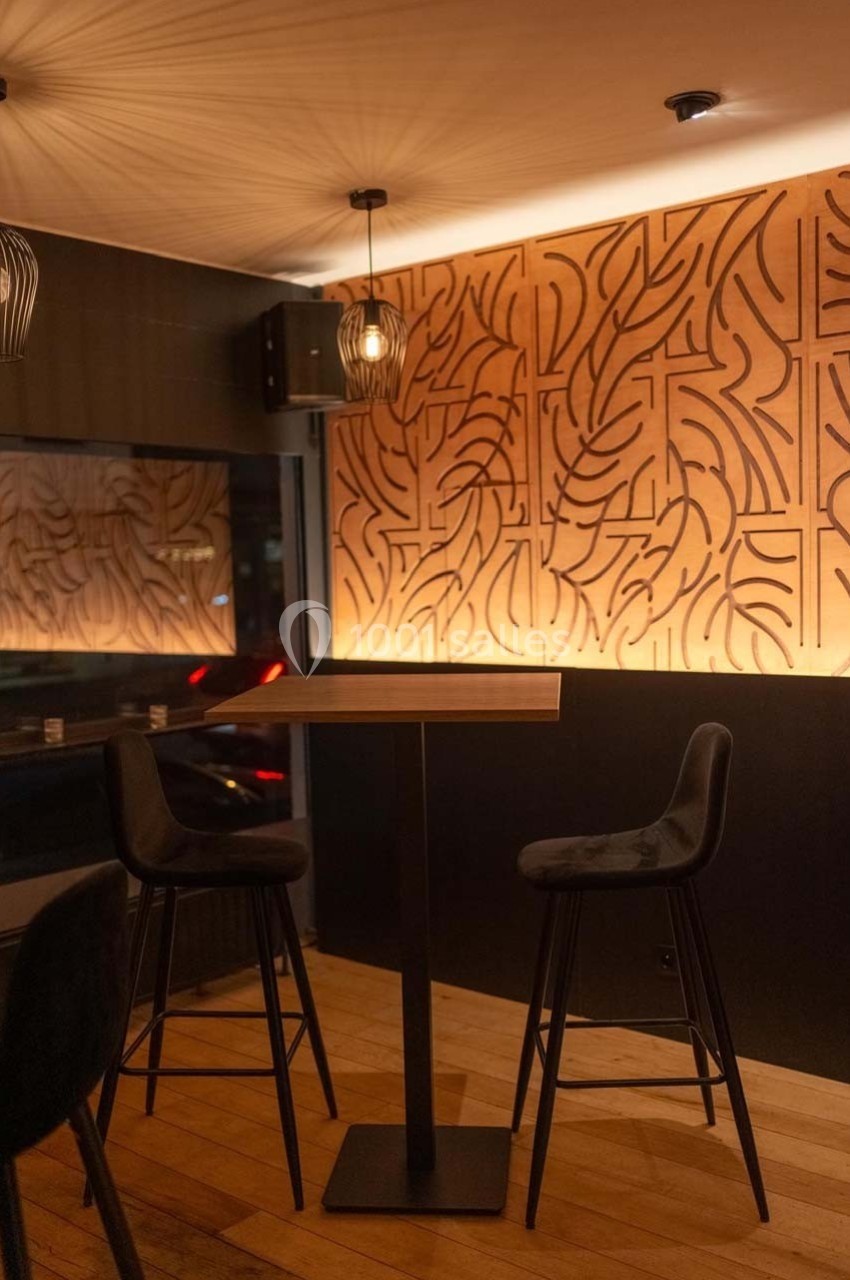 Table haute avec deux chaises noires dans un intérieur moderne, éclairé par des lampes suspendues et un mur décoratif en…