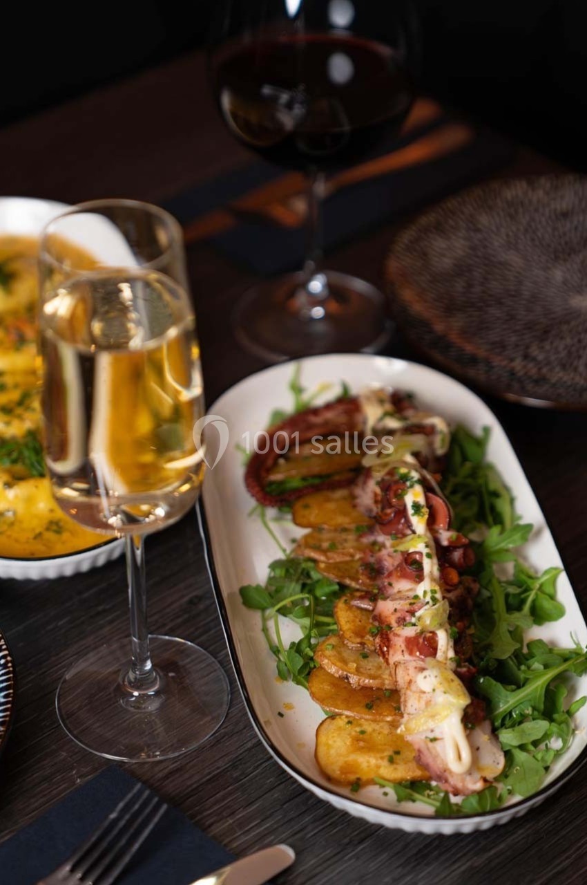 Assiette de poulpe grillé sur lit de roquette et pommes de terre, accompagnée de verres de vin et champagne.