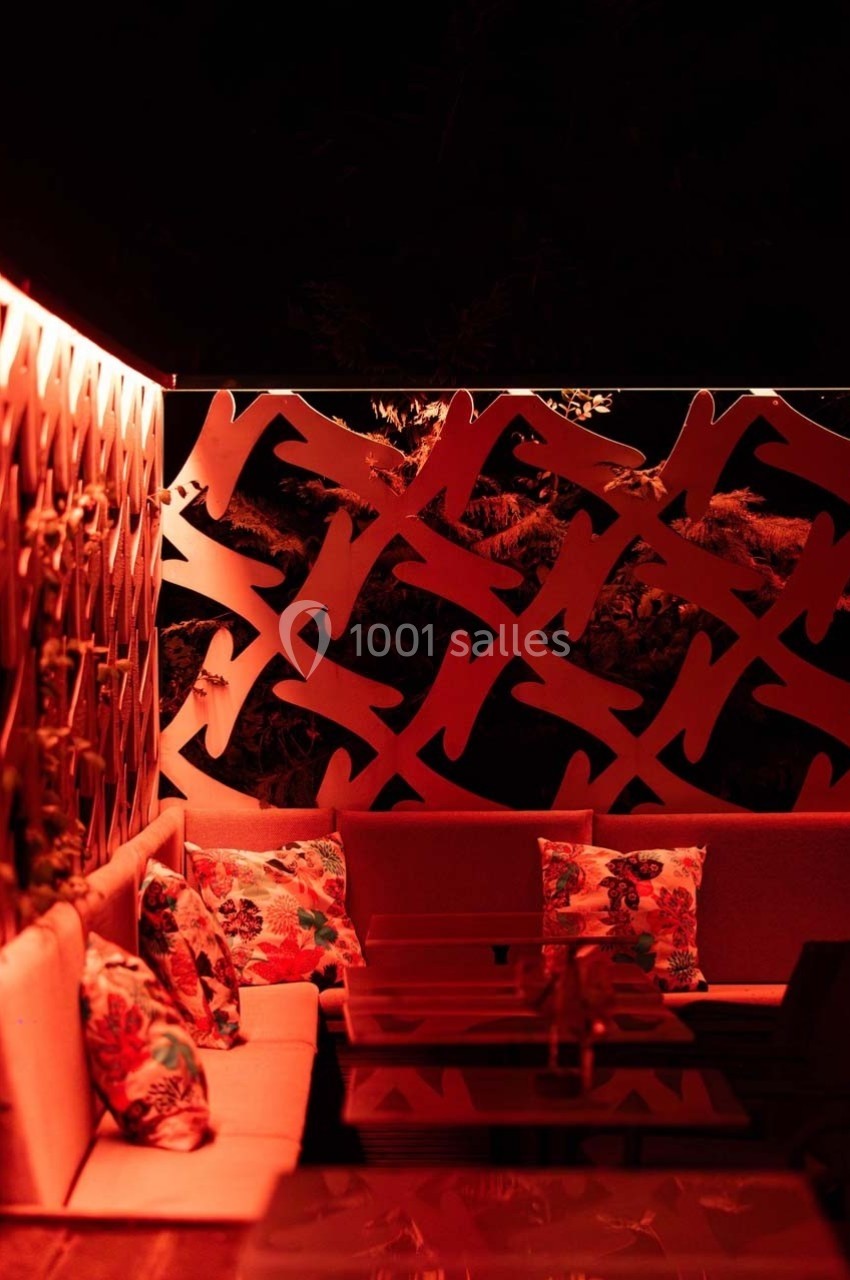 Espace extérieur éclairé de lumières rouges, avec banquettes, coussins à motifs floraux et cloison décorative ajourée.