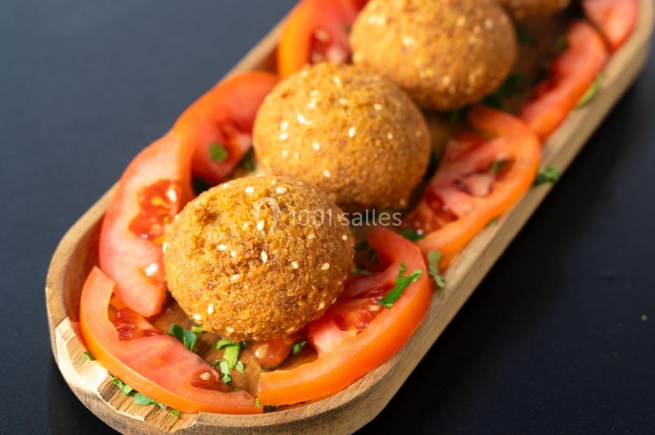 Assiette en bois contenant des falafels garnis de graines de sésame, posés sur des tranches de tomates fraîches.