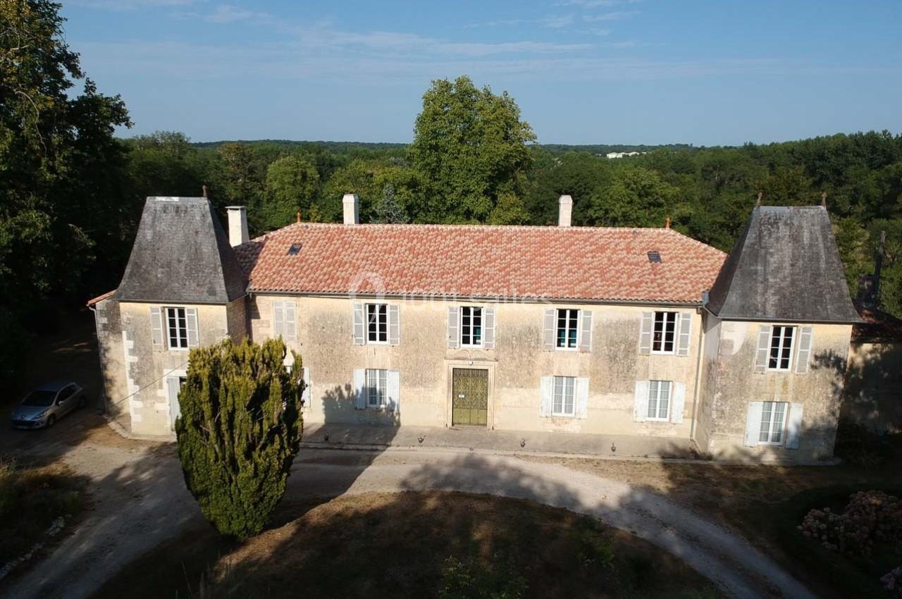 Façade d'un manoir ancien en pierre avec toit en tuiles, entouré de végétation et d'une allée gravillonnée.