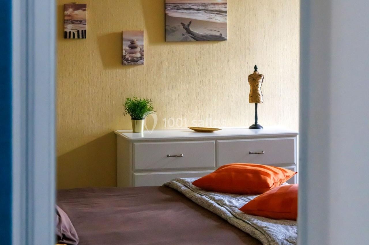 Vue d'une chambre avec un lit, des coussins orange, une commode blanche et des décorations murales.