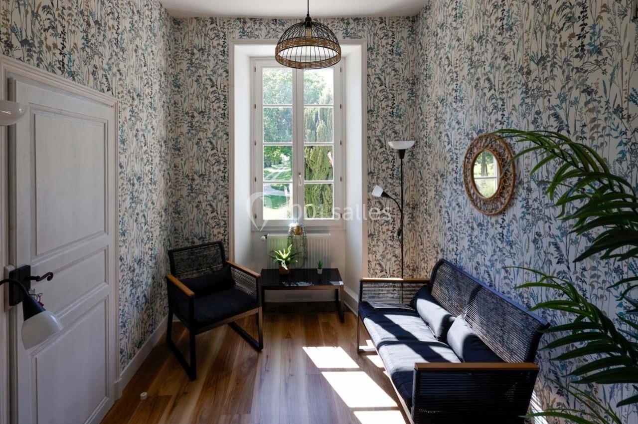 Petit salon lumineux avec murs tapissés de motifs floraux, mobilier en bois sombre et vue sur un jardin par une fenêtre.