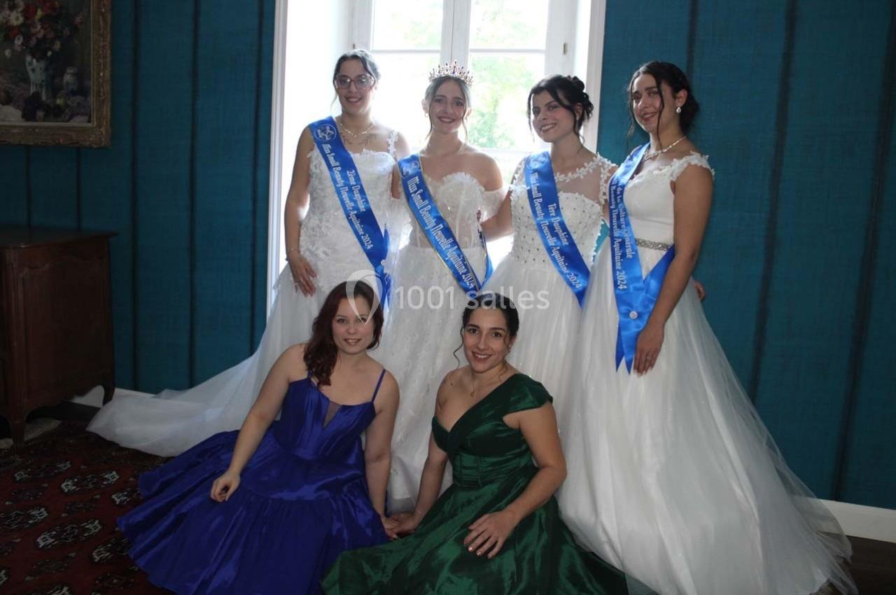 Six femmes en robes élégantes posent dans une pièce, quatre debout avec des écharpes et deux assises devant elles.