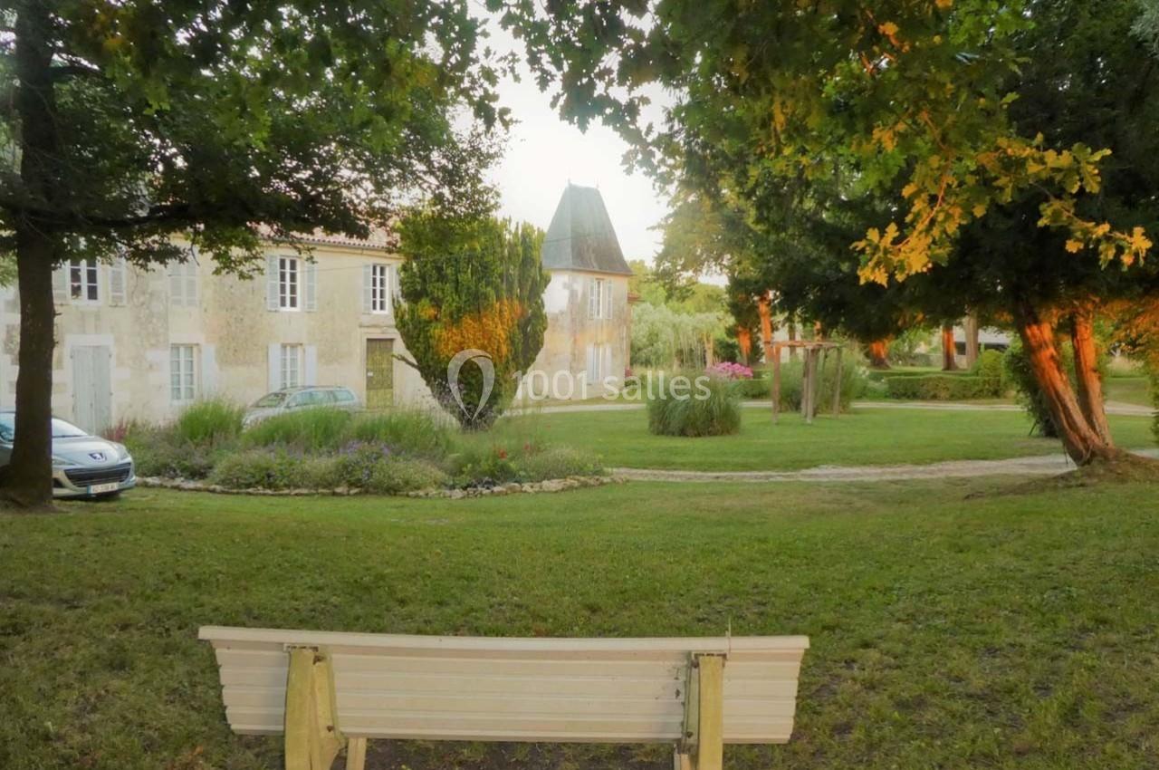 Banc en bois face à un jardin verdoyant avec une maison en pierre et des arbres au coucher du soleil.