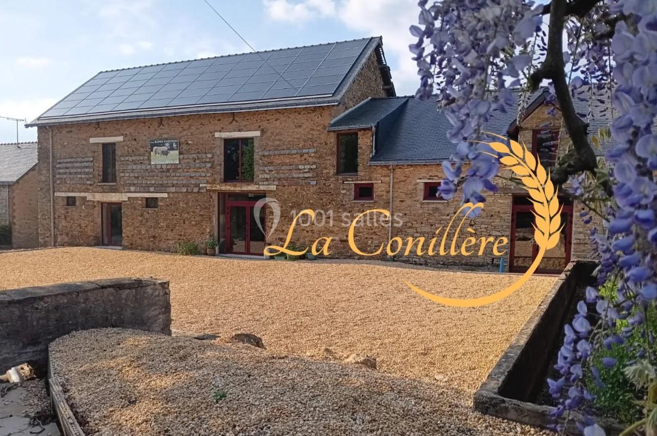 Bâtisse en pierre avec panneaux solaires sur le toit, cour en gravier et glycine en premier plan.