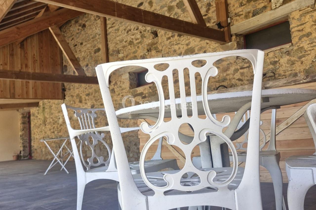 Chaises blanches en métal au design ajouré autour d'une table, dans une pièce avec murs en pierre et poutres apparentes.