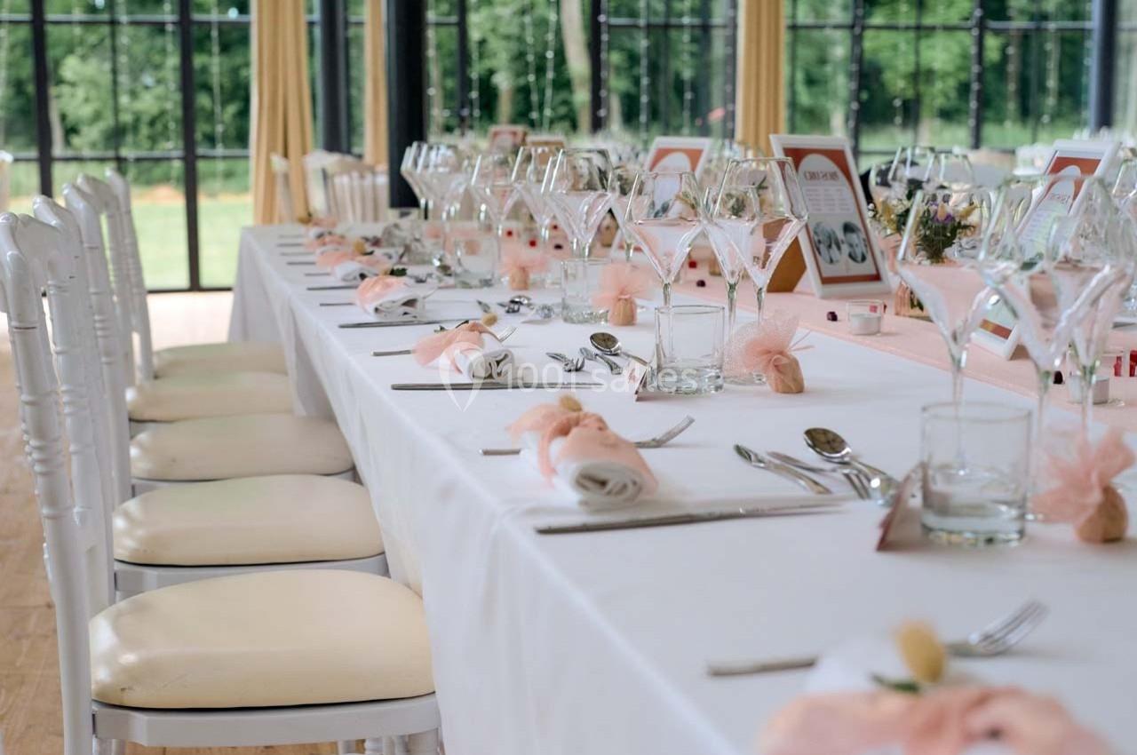 Table élégamment dressée avec nappes blanches, verres, couverts et décorations roses dans une salle lumineuse.