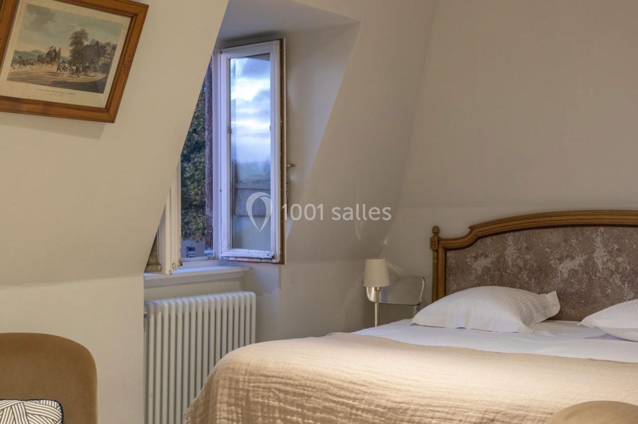 Chambre lumineuse avec un lit double, une fenêtre ouverte sur un paysage, un radiateur et un tableau au mur.