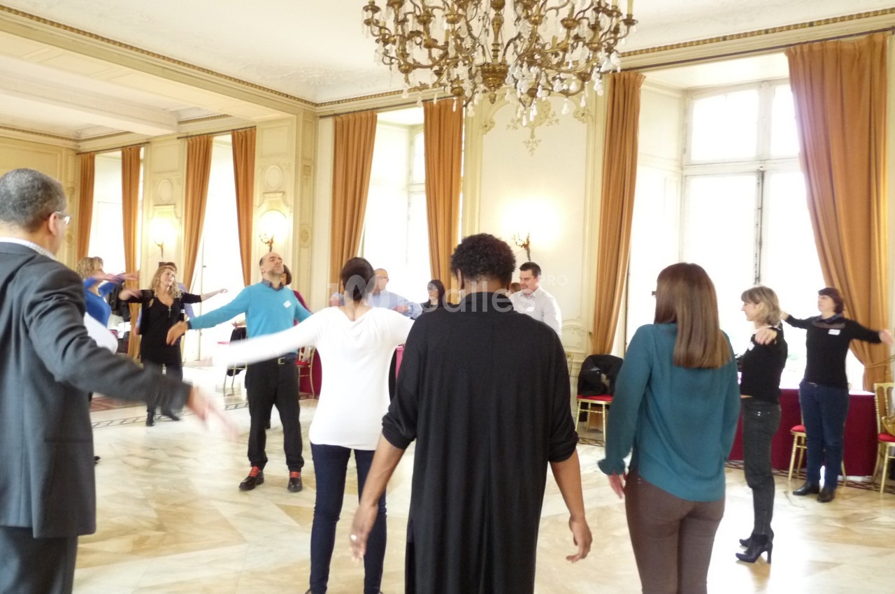 Des personnes debout en cercle dans une salle lumineuse avec un lustre, participant à une activité collective.