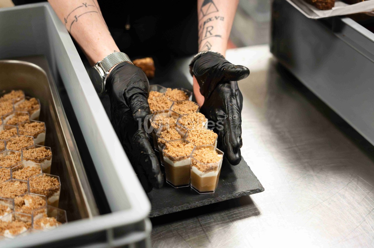 Une personne portant des gants noirs dispose des verrines garnies de crumble sur un plateau noir.