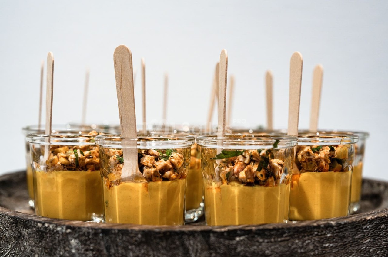 Verrines de soupe jaune garnies de morceaux croustillants et présentées avec des bâtonnets en bois.
