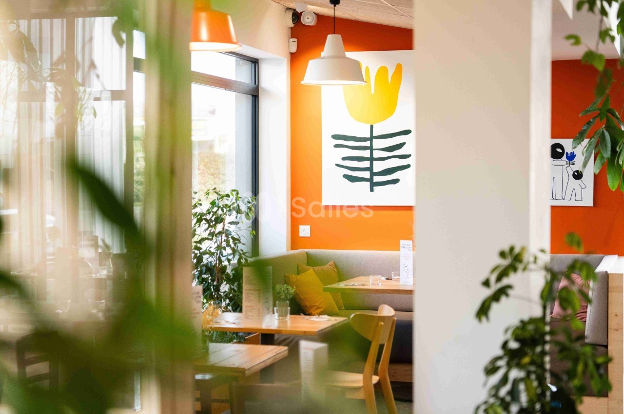 Intérieur lumineux d’un café moderne avec des plantes, des tables en bois et un mur orange décoré d’une peinture florale.