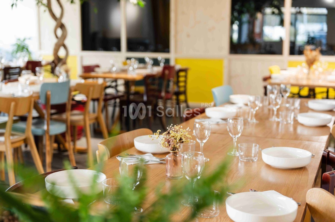 Salle de restaurant lumineuse avec tables en bois dressées, assiettes blanches et verres, décorée de plantes et fleurs…