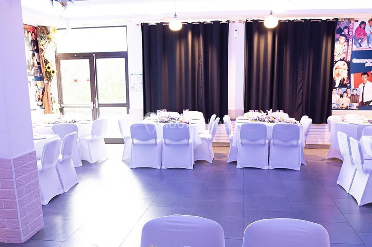 Salle de réception lumineuse avec des tables rondes dressées et des chaises blanches, rideaux noirs en arrière-plan.