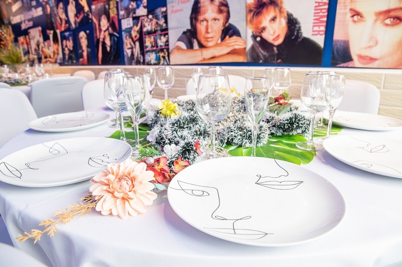 Table dressée avec assiettes décorées de visages stylisés, verres à pied, fleurs et centre de table végétal.