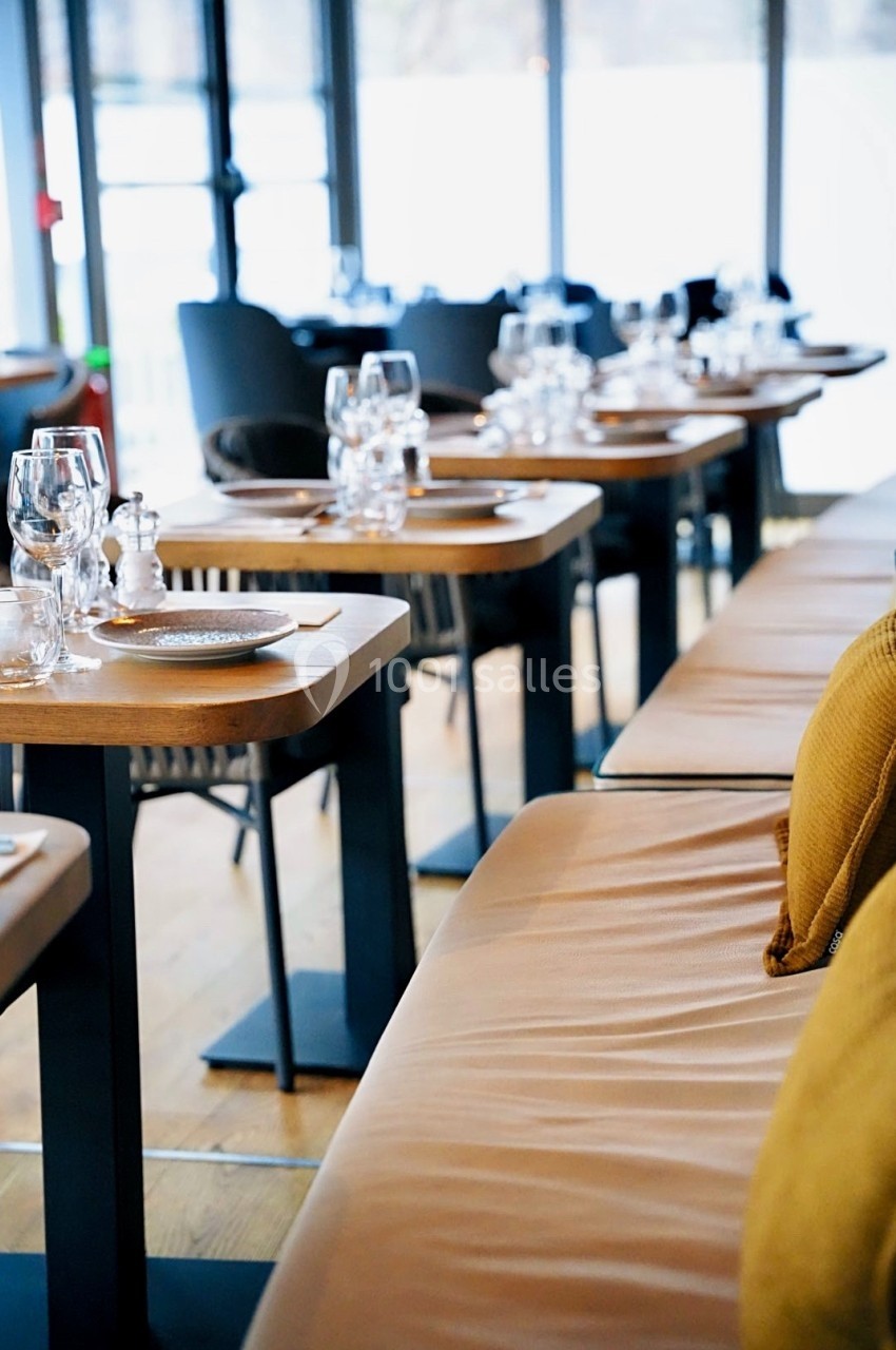 Tables en bois dressées avec verres et assiettes alignées dans un restaurant lumineux avec banquettes en cuir.