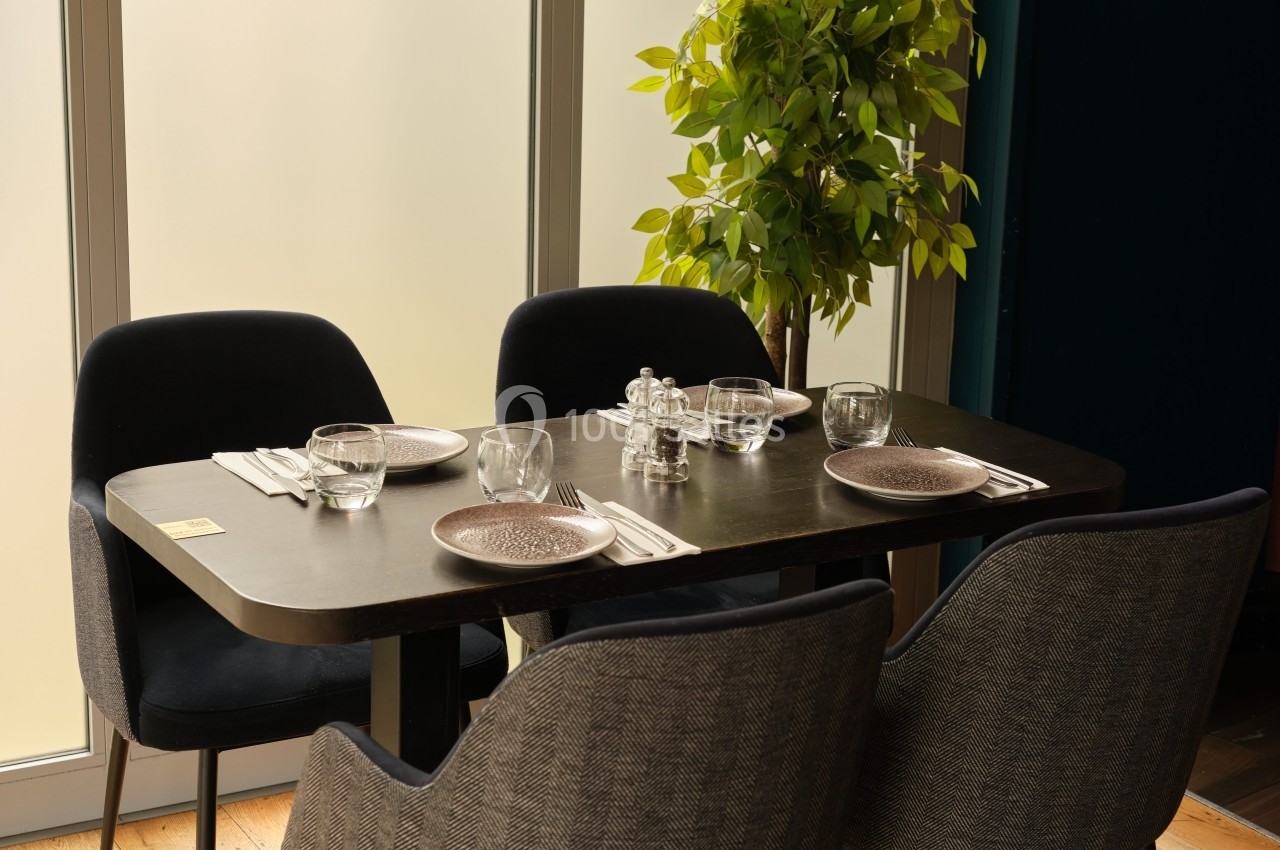 Table dressée pour quatre personnes avec assiettes, couverts et verres, près d'une plante et d'une baie vitrée.