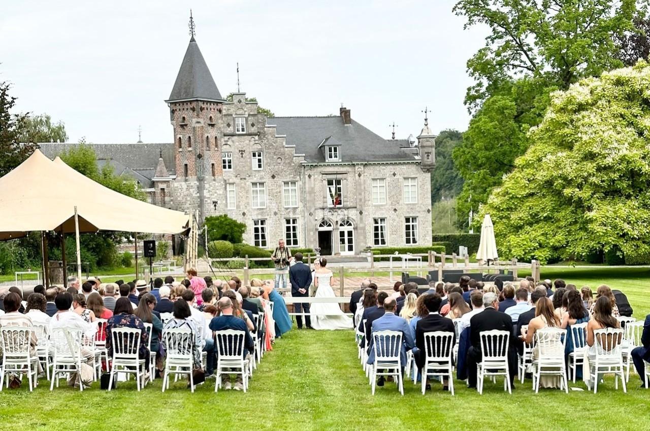 Cérémonie en plein air devant un château, avec des invités assis sur des chaises blanches alignées sur une pelouse.