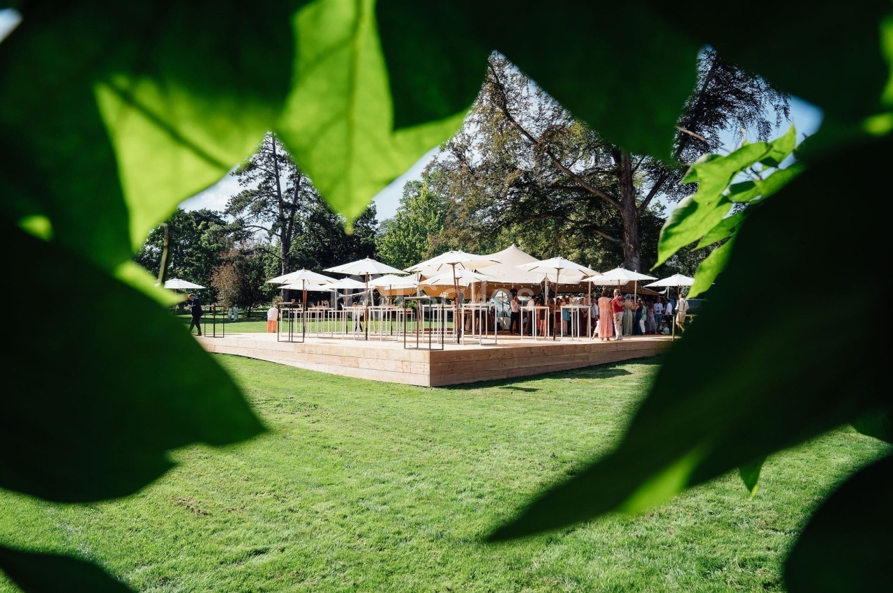 Terrasse en bois avec parasols blancs entourée de verdure, vue à travers des feuilles au premier plan.