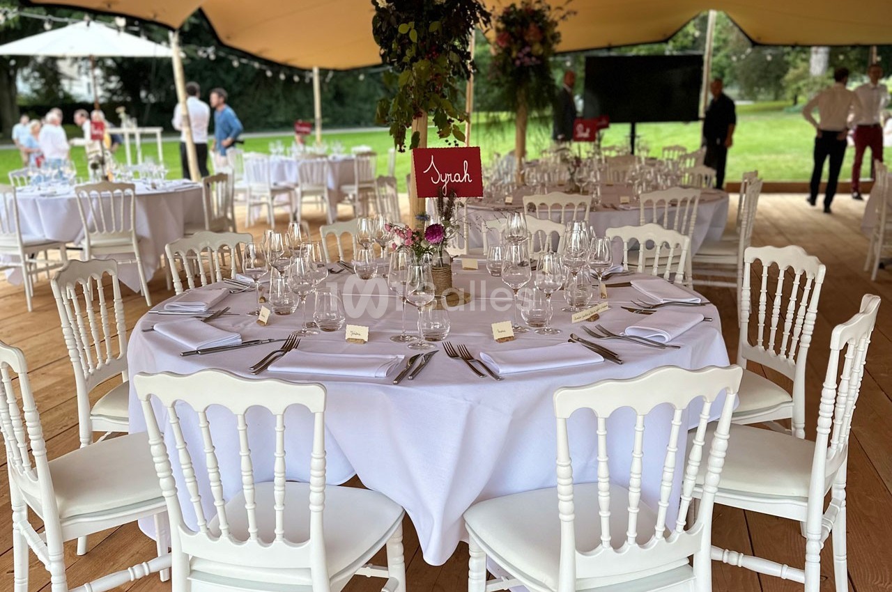 Table ronde élégamment dressée avec nappes blanches, chaises assorties et décoration florale sous une tente en extérieur.